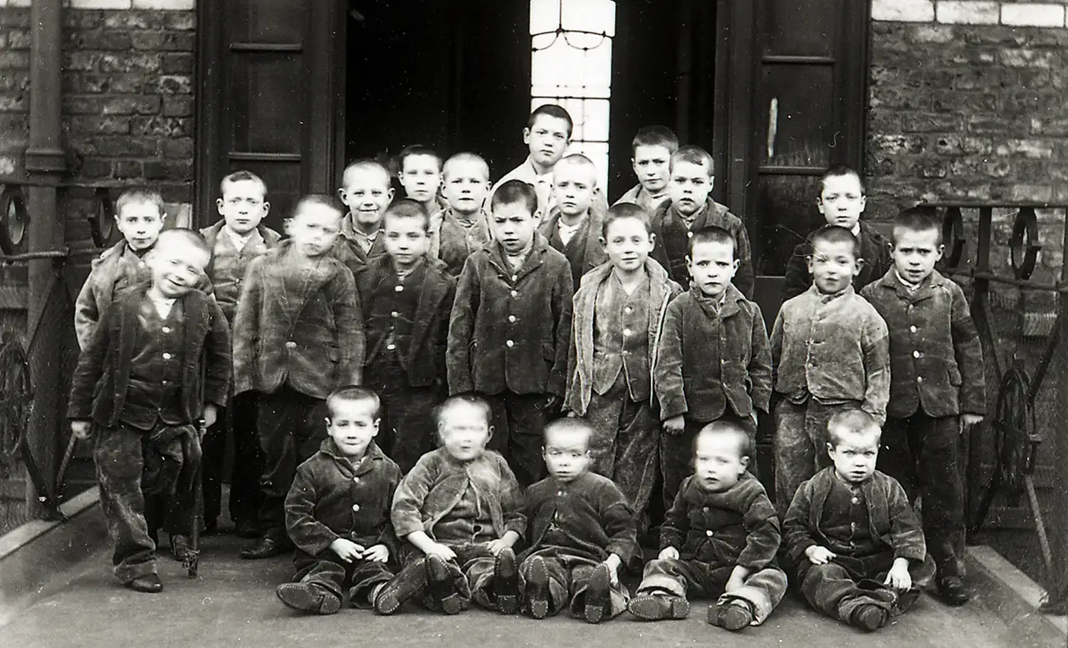 Twenty-three boys ranging from toddlers to about twelve years old are arranged for this group photograph. They all have their heads shaved and are wearing identical work clothes. The youngest sit in front, and one child appears to be missing a leg and using a cane.