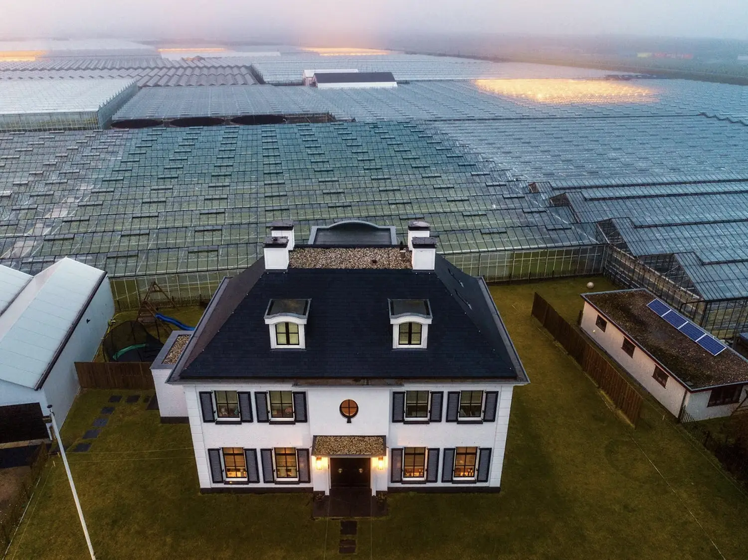 Greenhouses, some glowing with orange light, cover all ground visible in the image with the exception of where there stands a small traditional farmhouse.